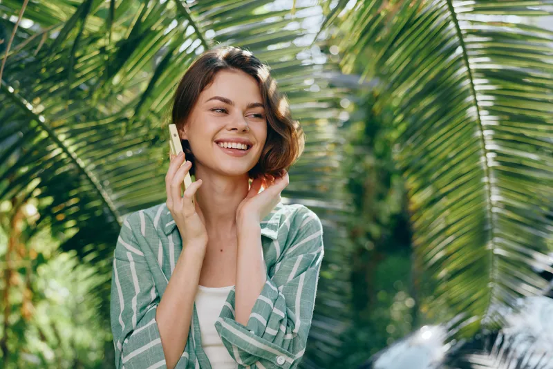 Cheerful woman outdoors, smiling while talking on a mobile phone, relaxed pose amid lush tropical plants and bright sunlight.
