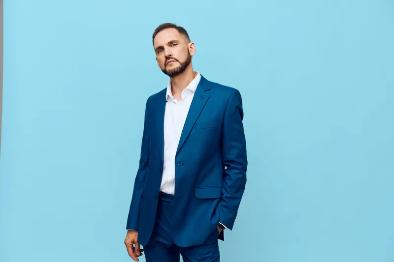 A composed business professional stands against a solid blue backdrop, projecting confidence and focus. His tailored suit communicates success while subtle emotion hints at determination and