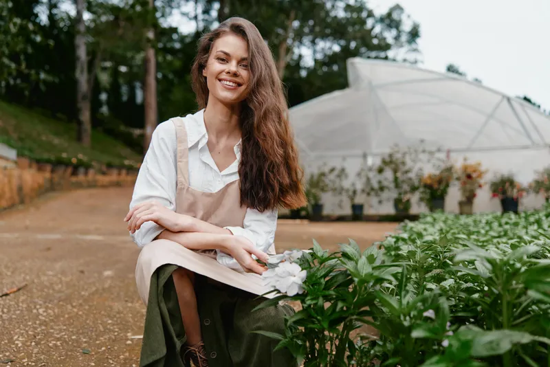 Smiling young woman in apron sitting in a greenhouse, surrounded by plants, embodies sustainable farming and healthy living ideals