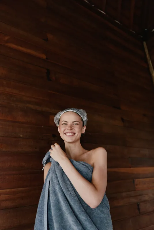 smiling woman wrapped in gray towel with hair cap enjoys wooden sauna interior, natural light highlighting warm atmosphere and relaxed mood.