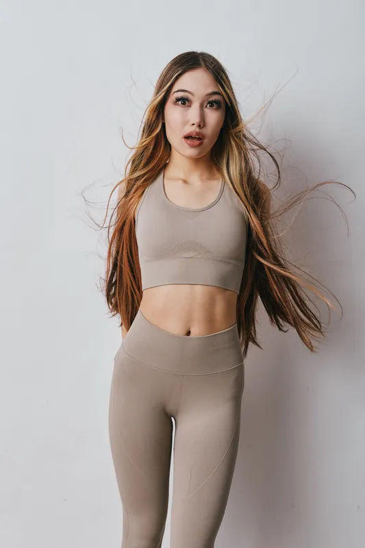 Young woman in beige seamless activewear poses with long hair and expressive face for fitness, yoga, wellness, health, workout and lifestyle studio portrait. Female model in sport outfit.