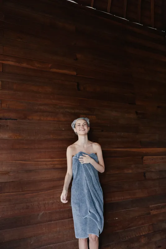 smiling woman wrapped in a blue towel standing against dark wooden wall, casual pose with towel on head, indoor spa or sauna atmosphere, natural lighting, relaxed mood