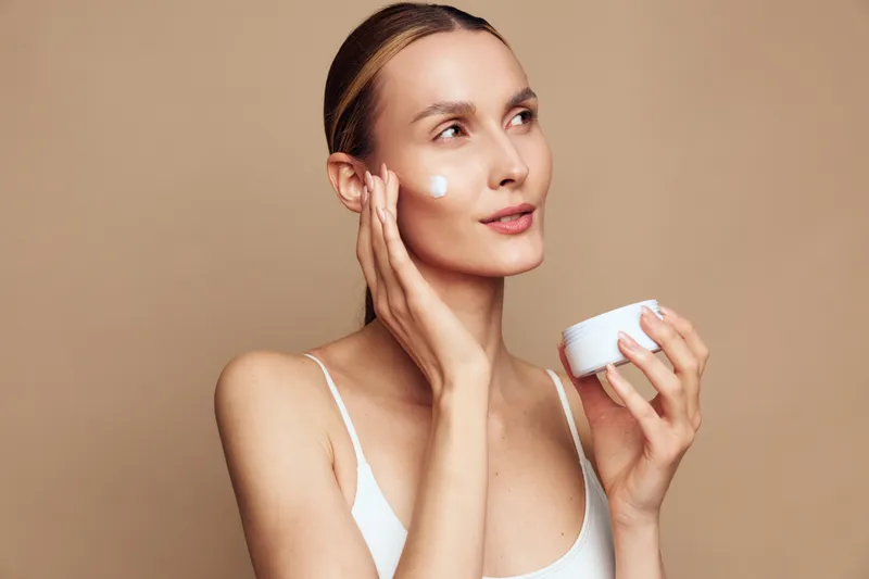 Woman applying skincare cream to face, holding small jar, using cotton pad, calm expression, smooth skin concept, minimal studio beige background, beauty care
