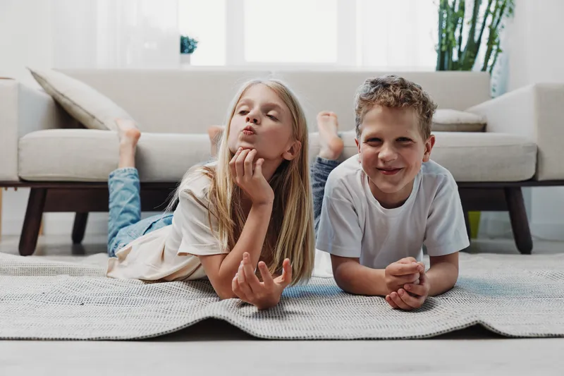 Two happy children lying on floor rug in bright living room, smiling and enjoying playful moment together at home interior.