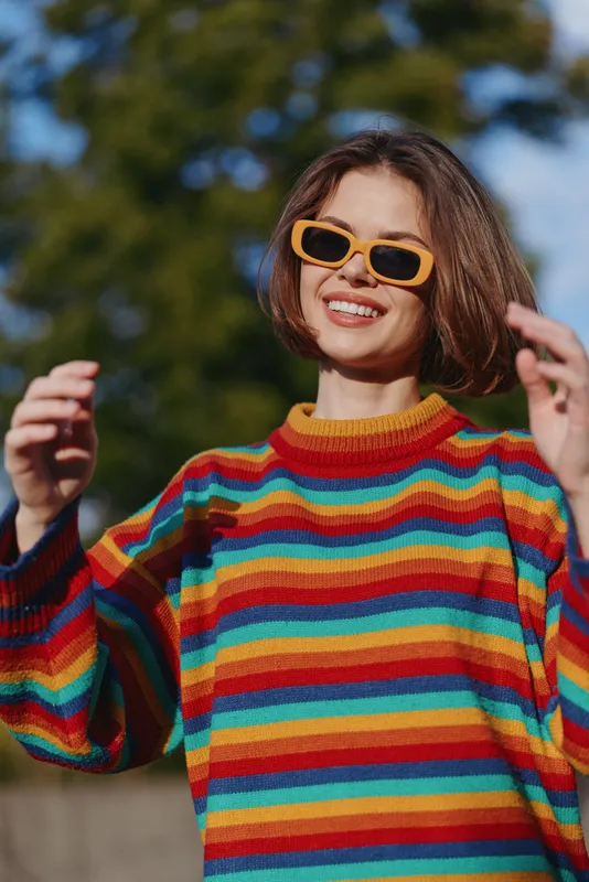 Young woman in a rainbow sweater and orange sunglasses smiling outdoors on a sunny day. Casual outfit, short hair, lifestyle portrait of joyful urban leisure and walking.