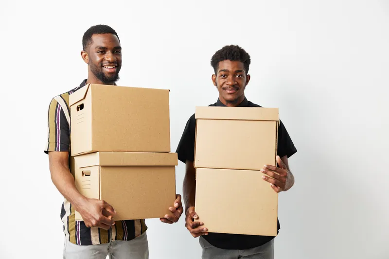 Happy black men holding cardboard boxes, smiling with enthusiasm, showcasing teamwork and collaboration during a move or organizational task.