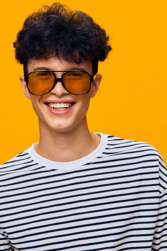smiling man wearing striped shirt and large yellow sunglasses against bright orange background, joyful young adult male with curly hair and casual style
