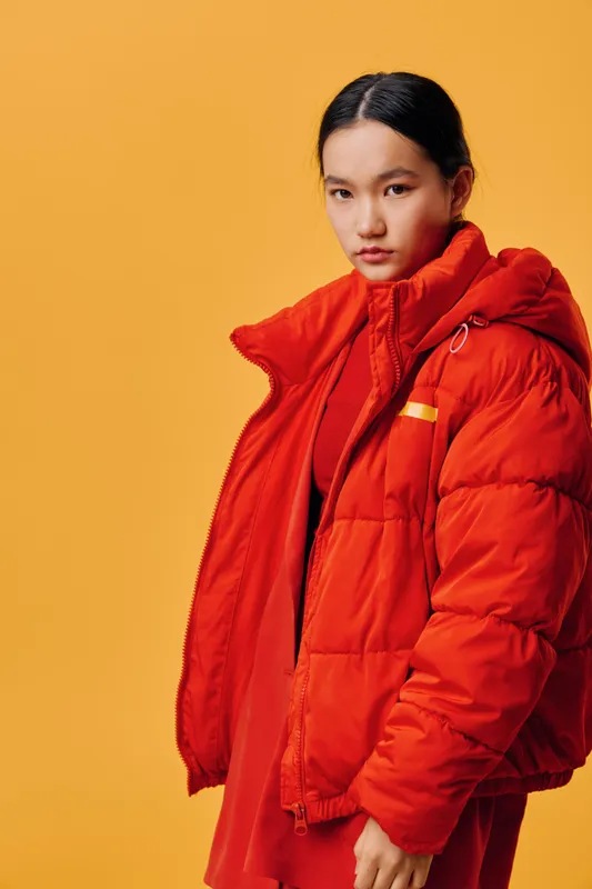 Young woman with long dark hair wearing a red puffer jacket and matching red dress standing against a solid orange background looking at camera with serious expression and side pose.