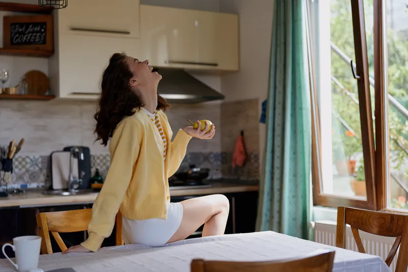 One young woman with curly hair wears a yellow cardigan and white shorts, seated on a kitchen table near a large window, laughing as sunlight fills the room with a warm, cheerful mood.