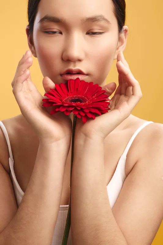 Asian woman gently holds a bright red flower near her face in a studio portrait with a warm amber backdrop, conveying calm, grace, and a contemplative mood.