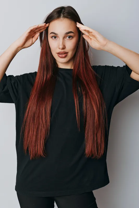 Young woman with long red hair and casual black shirt posing with hands near head on gray background. Female model with natural makeup and confident expression in studio portrait.