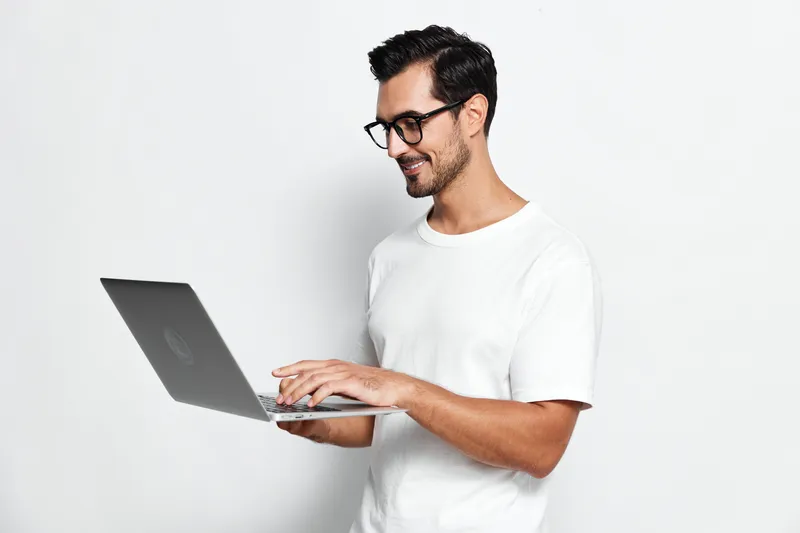 Smiling man with glasses working on laptop, standing against plain solid white background. Engaging in technology and modern lifestyle concept.