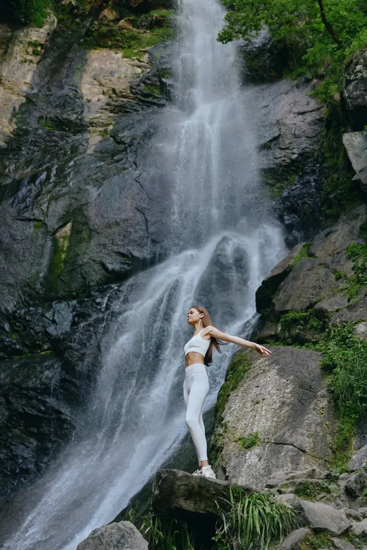 woman standing near waterfall in white fitness clothing with arms outstretched, surrounded by lush green nature and rocky terrain, expressing freedom and balance