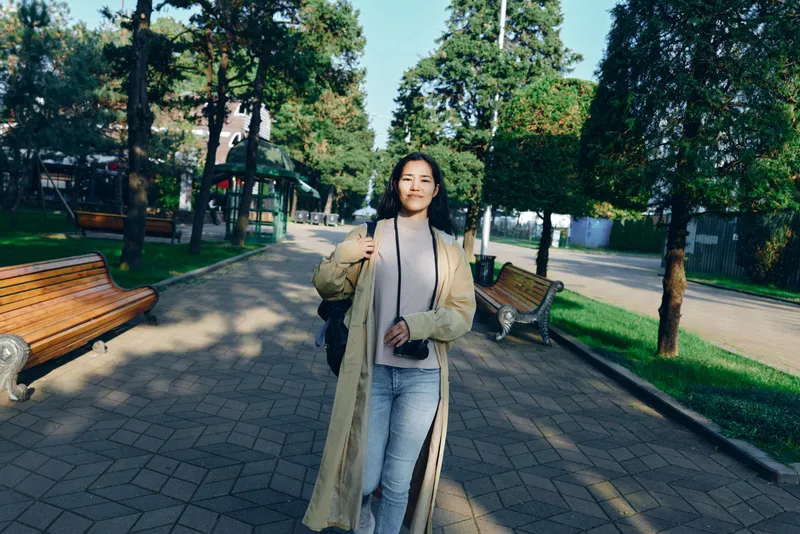 Woman in a long beige coat walks along a shaded park path, carrying a camera over her shoulder, casual attire, confident stride amid trees, benches, and a sunny urban park setting