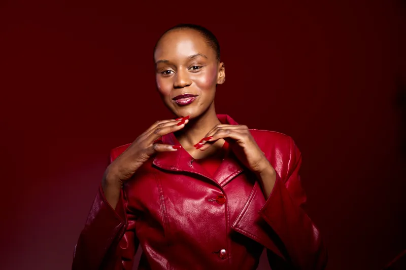 Confident dark skinned African woman wearing shiny red leather jacket poses smiling with hands near face on dark red background. Concept of fashion, beauty, and elegance in studio.