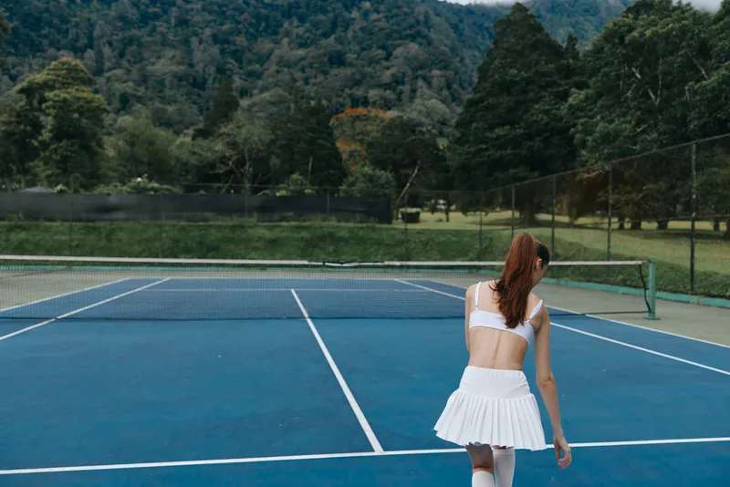 Young female tennis player in sportswear on blue court, showcasing focus and determination amid lush green background, perfect for sports and fitness themes