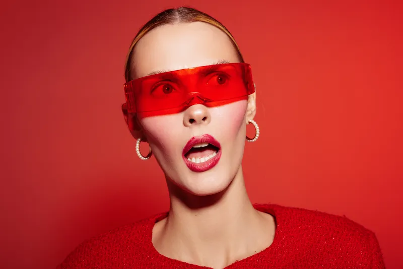 Surprised woman with red lips and red sunglasses looks shocked on red background. Fashion portrait with bold makeup and modern accessories in studio lighting.