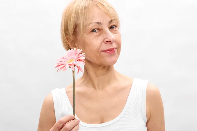 Elderly woman portrait holding a pink flower, smiling gently in a bright studio with white background, calm mood, spring freshness, soft focus concept for aging and wellbeing.
