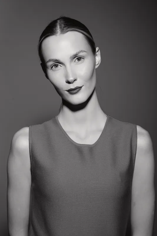 Portrait of a slim Caucasian woman with pale skin and slicked back hair wearing a sleeveless top, black and white studio photo.