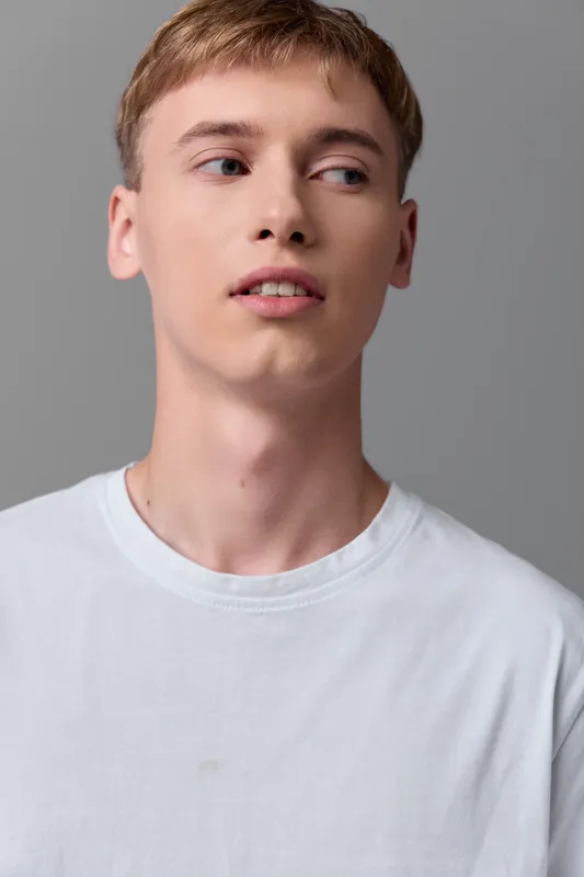 Young man in a white shirt stands against a neutral studio backdrop, conveying a thoughtful, casual mood suitable for fashion and lifestyle campaigns, clean minimal composition, versatile for