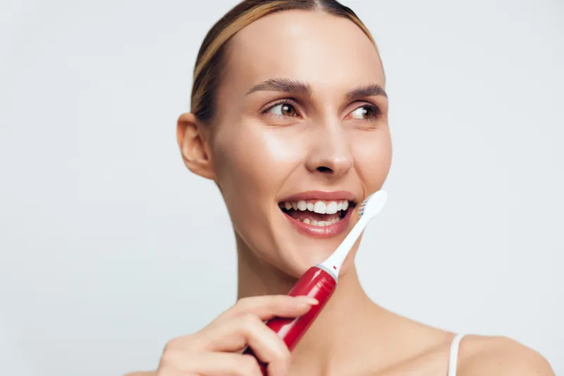 Young woman with clear skin smiles while holding electric toothbrush near mouth isolated on white background, demonstrating dental hygiene and oral care routine.