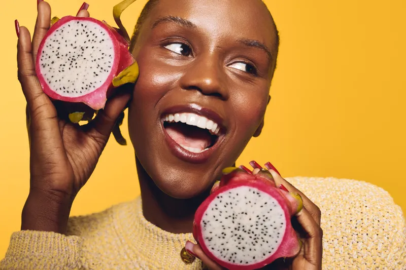 Smiling African woman with dark skin holds two halves of dragon fruit near her face on yellow background. Concept of healthy lifestyle, tropical fruit, joy, and natural beauty.