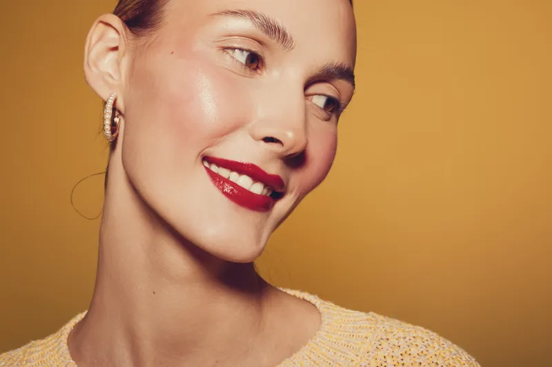 Close-up of smiling woman with red lipstick and glowing skin wearing earrings and a light sweater on warm yellow background. Concept of beauty, makeup, and skincare.