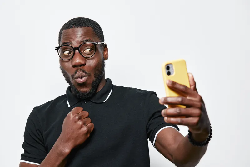 Black man with dark skin and glasses taking selfie with yellow smartphone, making surprised face and fist gesture in casual black polo shirt on white background.