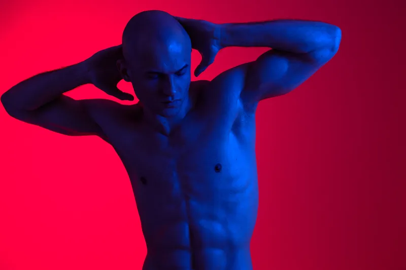 A striking image of a man posing dramatically against a vibrant red background, showcasing his toned physique and unique color lighting effects.