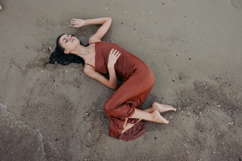 Relaxed woman lying on beach sand wearing a flowing brown dress, with closed eyes and calm expression, embodying peace and nature connection.
