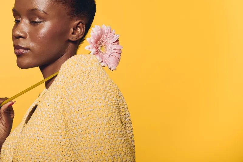 Portrait of an African woman with dark skin holding a pink flower behind her neck wearing a yellow textured sweater on a yellow background. Concept of natural beauty and fashion.