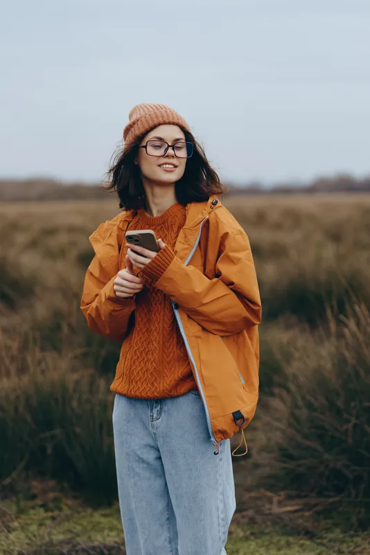 Woman lifestyle outdoors autumn field, orange jacket and knit hat, smiling with glasses, holding smartphone, casual walk, soft cloudy light, natural colors, calm mood