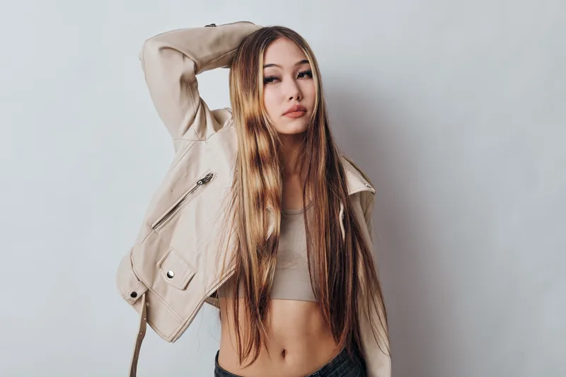 Young woman with long hair wearing casual light jacket and crop top posing against plain wall. Female model standing with hand on head in studio, fashion and beauty portrait.