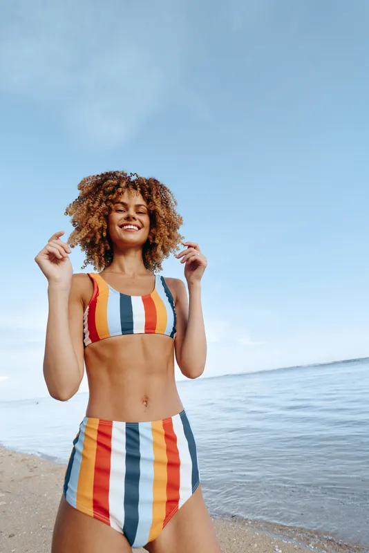 A smiling young woman in a colorful striped bikini stands on a sunny beach, posing confidently. Clear blue sky, calm sea, light breeze, carefree mood, summer vibe, woman enjoying vacation outdoors.