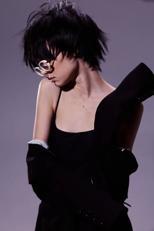 Stylish woman with short black hair wearing white glasses and black outfit posing with elegance in a minimalistic studio setting.