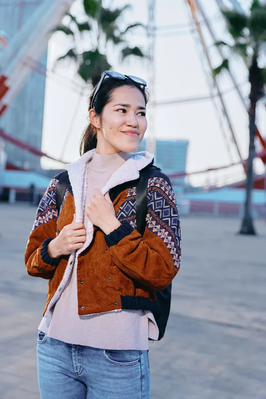 Smiling woman in layered autumn jacket and scarf stands outdoors, backpack on shoulder, urban plaza with palm trees, casual fashion and travel vibe captured in daylight