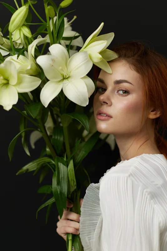 Young woman with long auburn hair holding white lilies against a dark background, conveying elegance and tranquility in a serene moment