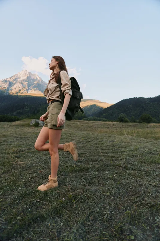 A young woman with a backpack hikes through a wide grassy valley, mountains in the background. Focus on outdoor adventure, wellness, and nature.