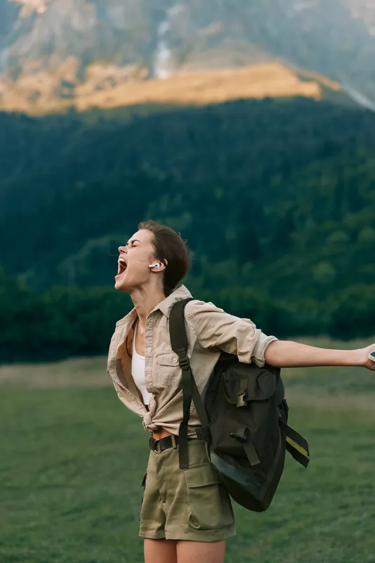 Lively woman outdoors, laughing with mouth open, backpack on shoulder, wide grassy field, mountains in the background, sunlit moment, carefree lifestyle