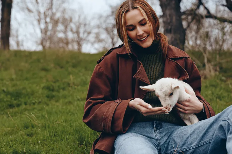 A woman in a brown coat sits on the grass outdoors, gently holding a goat kid, creating a calm moment of care and affection in a pastoral field that evokes everyday lifestyle and connection with