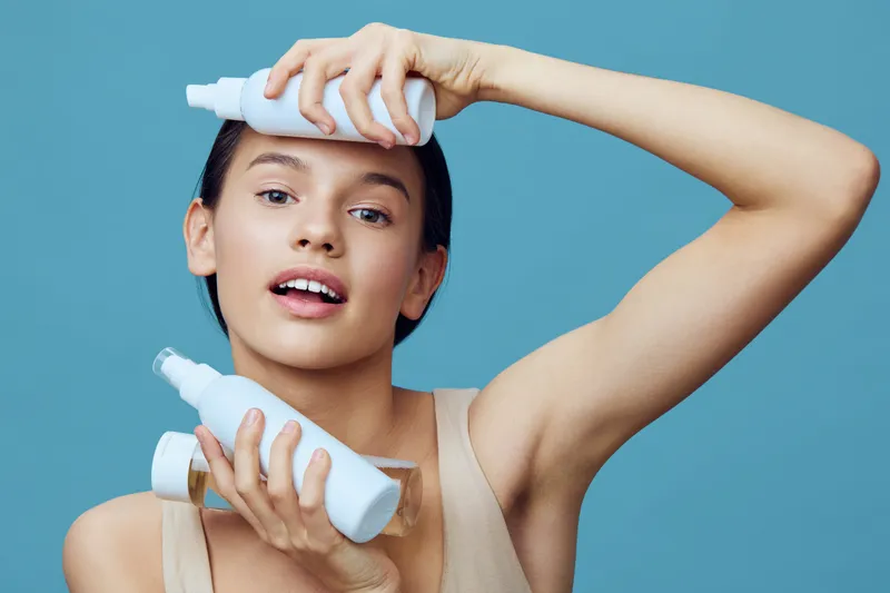 Young woman holding skincare products with a fresh expression, showcasing a beauty routine against a soft blue background, emphasizing healthy skin and self care