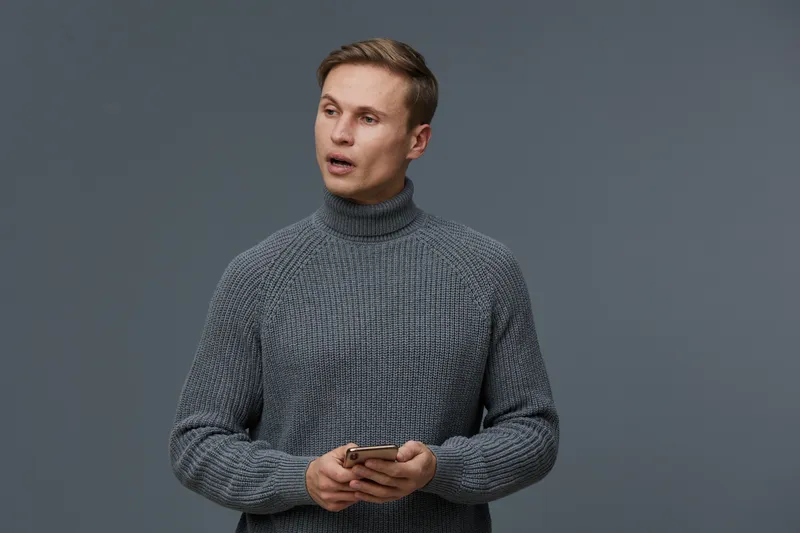 Man with short brown hair wearing gray turtleneck sweater holding smartphone, standing against plain gray background. Casual indoor portrait with focused expression, suitable for technology, communica