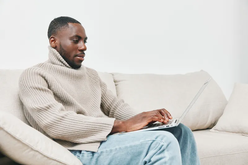 Young Black man in cozy sweater working on laptop at home, showcasing focus and comfort in a modern living space