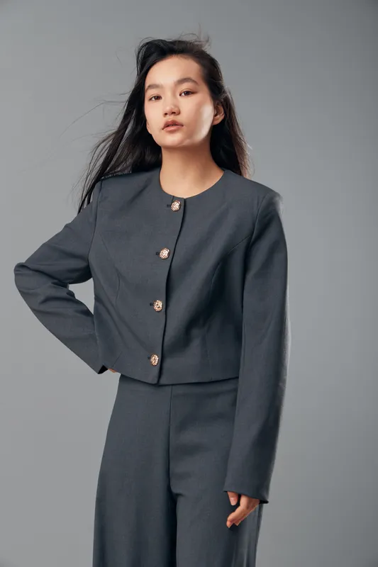 Young woman wearing gray formal suit standing with hand on hip against neutral background, looking confidently at camera with natural makeup and long dark hair.
