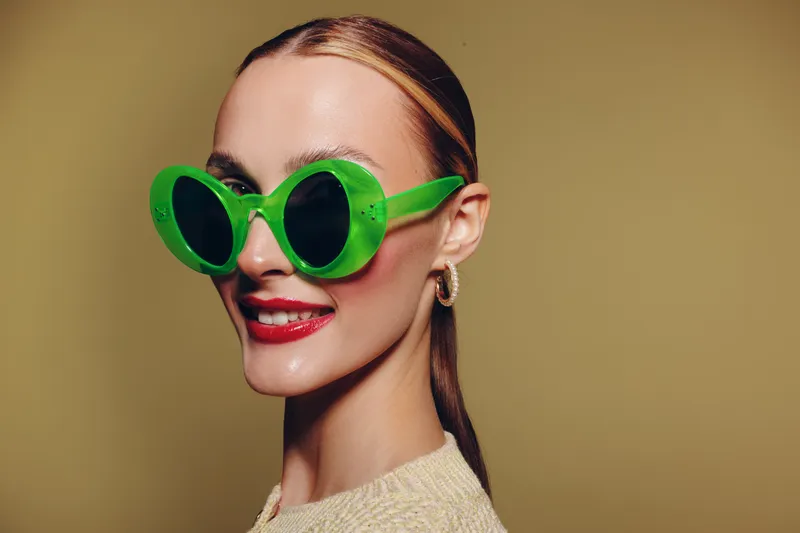 Young woman with slicked back hair wearing large green sunglasses and red lipstick smiles confidently against a beige background. Concept of fashion, style, and beauty portrait.