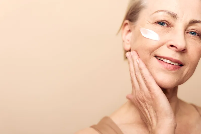 Mature woman with clear skin applies cream on cheek smiling gently against beige background. Concept of skincare, aging beauty, self-care, and natural wellness for healthy complexion.