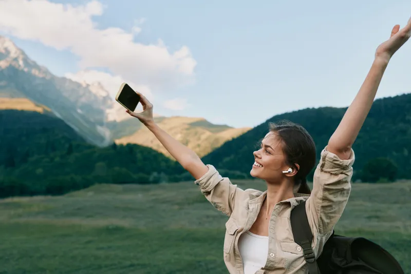 Young woman with earbuds raises her arms joyfully while taking a selfie with a smartphone in a mountainous outdoor setting during daytime.