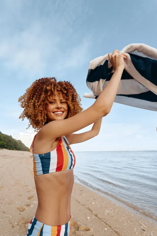 Young woman with curly hair joyfully raises a striped towel above her head while standing on a sandy beach near the calm sea under a clear sky.