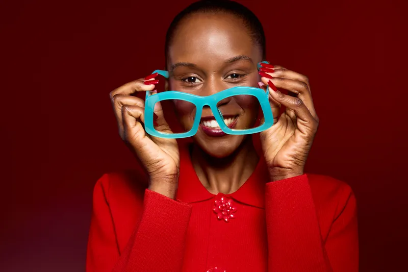 Smiling African woman with dark skin holds large blue glasses in front of her face wearing red sweater. Bright red background and cheerful mood create concept of fashion and joy.