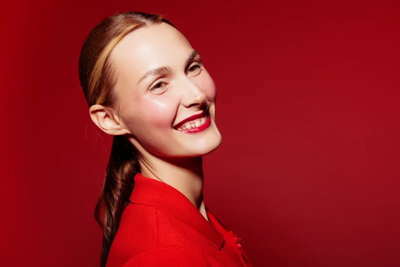 Smiling woman with red hair and red lipstick wears red sweater against red background. Portrait of happy female model with natural makeup and bright studio lighting.
